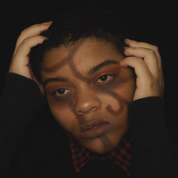 a black man with short hair, round black earrings, and a septum ring. he has brown twisting vines on his face. he holds his face