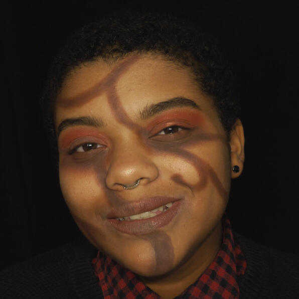 a black man with short hair, round black earrings, and a septum ring. he has brown twisting vines on his face. he smiles.
