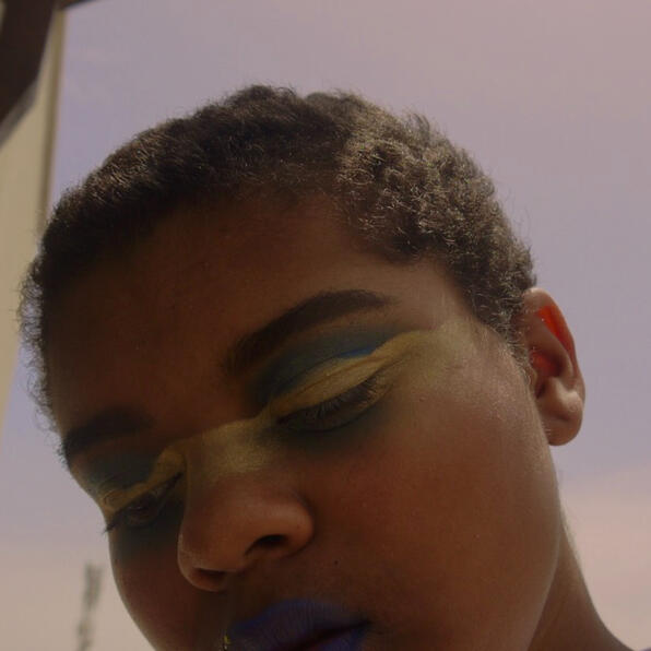 a black man with blue and gold clouds on his eyes and blue and gold on his lips. he tilts his head down in an endless sky.