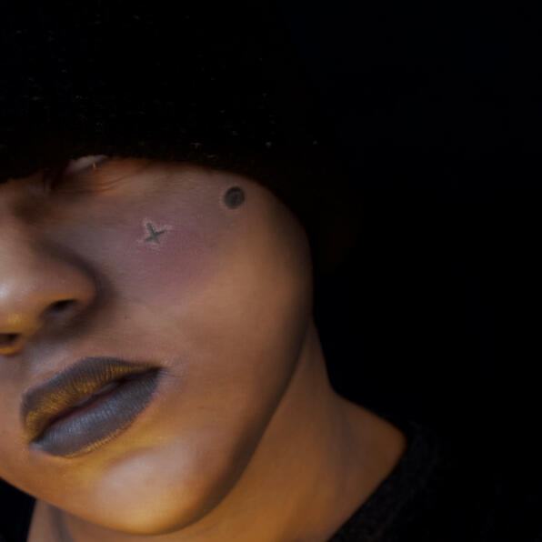 a black man shrouded in darkness, a dark galaxy painted across his cheeks. his eyes are white, half his face obscured.