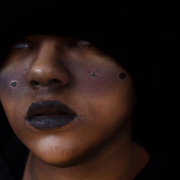 a black man shrouded in darkness, a dark galaxy painted across his cheeks. his eyes are white, lifting into the black void.