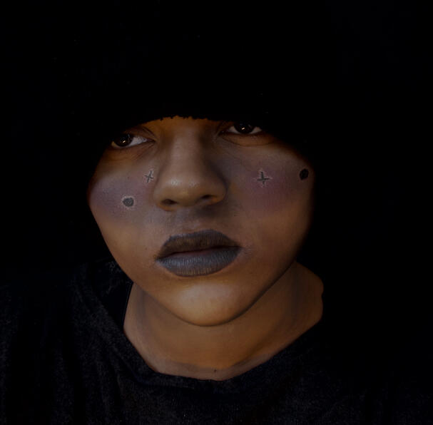a black man shrouded in darkness, a dark galaxy painted across his cheeks. his eyes are dark and open.