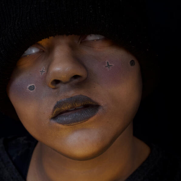 a black man shrouded in darkness, a dark galaxy painted across his cheeks. his eyes are white, staring into you.