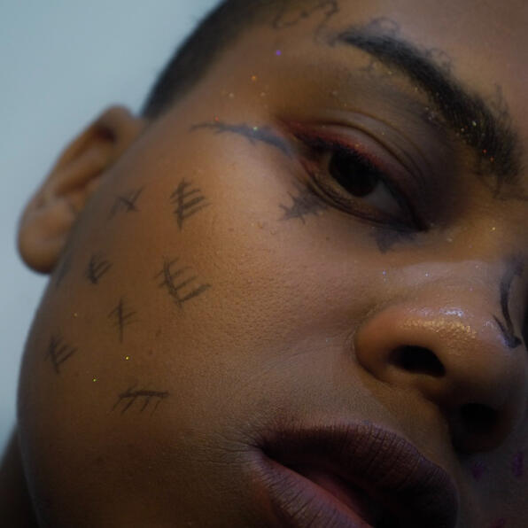 a black man with barbed wire for eyebrows, spikes under eye, and tally marks on his cheek. he looks onward.